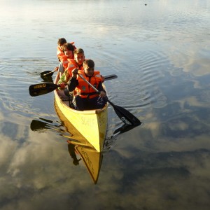 Vier Kinder mit Schwimmweste padeln mit einem gelben Kanu über den See. Inklusive Sommerreise des Indiwi Berlins.